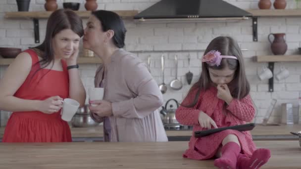 Mormor, mor och lite daghter är tillsammans i köket. Den flicka som leker på tabletten sitter på bord, mormor viskar hemlighet på mor öra, båda leende. Begreppet generationer. — Stockvideo