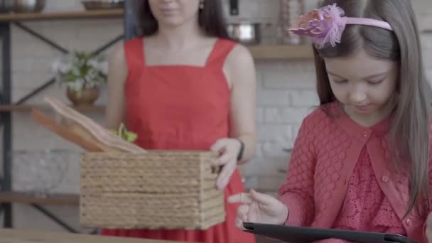 Молода мати поклала на стіл коробку з хлібом і розмовляла зі своєю маленькою донькою, яка грає в ігри на планшеті . — стокове відео