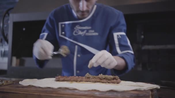 Skillful man in blue chef uniform cooking lula kebab in modern restaurant. Cook puts topping on the grilled minced meat lying on pita lavash close up — Stock Video