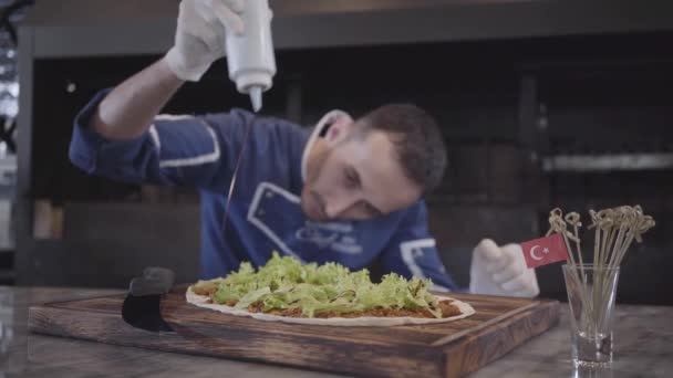 Homme habile en uniforme de cuisinier préparant un plat savoureux enveloppé dans de la lavande. Chef en gants de caoutchouc blanc versant des feuilles de laitue gisant sur pita avec sauce barbecue de la bouteille. Cuisine turque — Video