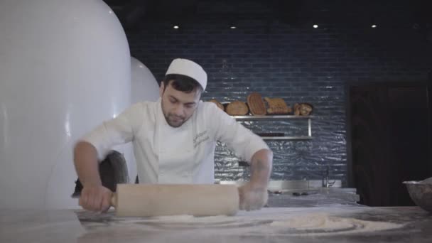 Chef en uniforme blanco trabajando masa rodante rápida con gran rodillo de masa de madera en la mesa en la cocina moderna del restaurante. El horno de leña está en el fondo. Concepto de preparación de alimentos — Vídeos de Stock