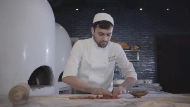 Chef in white uniform working fast putting raw cutted meat on the dough base lying on the table in modern restaurant kitchen making fatayer. Big oven is in the backgound. Turkish food preparation — Stock Video