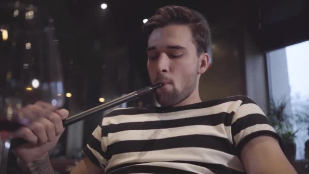 Hombre guapo disfrutando de la cachimba sentado en el moderno bar restaurante cafetería. Cliente seguro exhala nubes de vapor sentado en un hookah de cerca. Hombre guapo relajándose solo — Vídeos de Stock