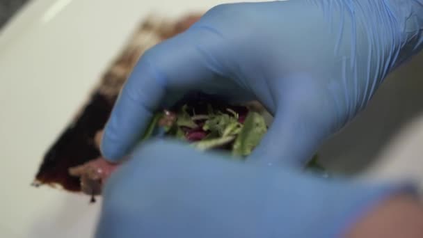 Les mains de chef dans des gants de caoutchouc bleu mettre la salade près du morceau de viande marinée crue sur l'assiette fermer dans le restaurant moderne. Cuire en préparant des aliments de viande. Mode de vie sain — Video