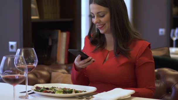 エレガントな笑顔の女性はモダンなレストランで座っています。現代の女性は、高級レストランで彼女の食べ物の写真を取る。おいしい料理を食べて快適な場所で時間を過ごす女性 — ストック動画