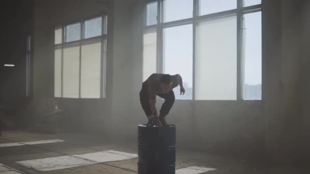 Hombre caucásico joven hábil con tatuaje en la mano delante de una gran ventana en la oscuridad en el edificio abandonado salta del barril de gas y se aleja. Movimiento lento — Vídeo de stock