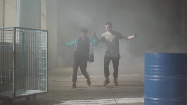 Dos jóvenes bailando en la oscura y polvorienta habitación del edificio abandonado. Adolescentes haciendo movimiento de baile simultáneamente, tomados de la mano, practicando en break-dance. Movimiento lento — Vídeo de stock