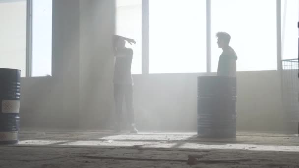 Dos jóvenes con ropa casual en el oscuro y polvoriento edificio abandonado practicando el baile frente a la ventana.. Un adolescente haciendo movimientos de baile, su amigo mirándolo. Chicos abrazos, terminando con éxito — Vídeos de Stock