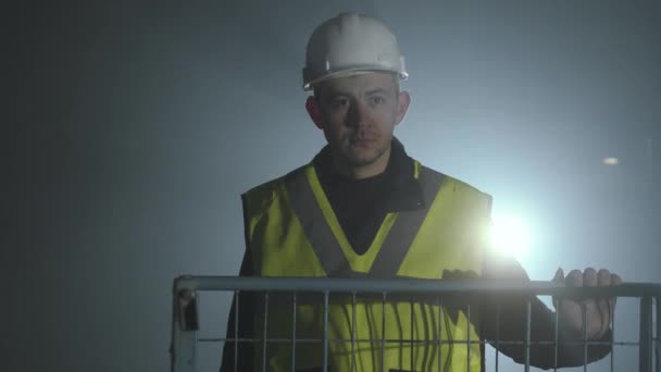 Серйозна людина в формі будівельників і шолом, що стоїть перед чорним тлом з прожектором. Портрет вдумливого архітектора з рукою на клітці. Студійний знімок — стокове відео