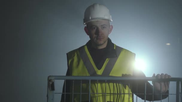 Uomo serio in uniforme costruttori e casco in piedi davanti allo sfondo nero con riflettori. Ritratto di architetto premuroso con la mano sulla gabbia. Studio girato — Video Stock