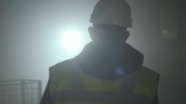 Вид ззаду на людину у формі будівельників і шолом, що йде перед чорним тлом з прожектором. Портрет вдумливого архітектора, який виконує свою роботу. Студійний знімок — стокове відео