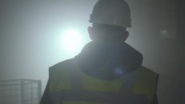 Vue arrière de l'homme dans l'uniforme des constructeurs et casque marchant devant le fond noir avec projecteur. Portrait d'architecte attentionné faisant son travail. Plan studio — Video