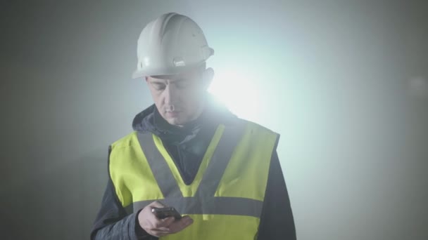 Porträt des Mannes in Bauarbeiter-Uniform und Helm-SMS auf dem Handy vor schwarzem Hintergrund mit Scheinwerfer. Porträt eines nachdenklichen Architekten, der seine Arbeit macht. Studioaufnahme — Stockvideo