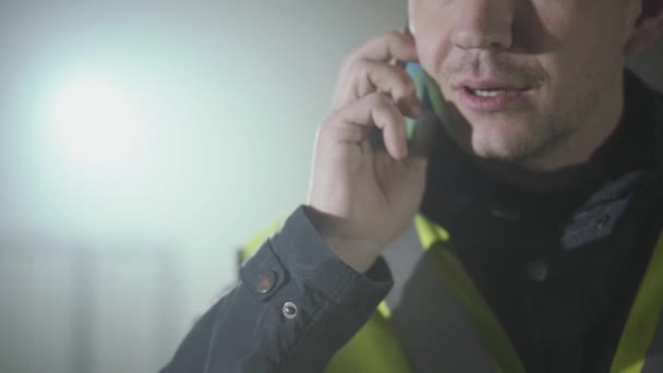 Närbild porträtt av mannen i byggare uniform och hjälm pratar med mobil telefon framför den svarta bakgrunden med Spotlight. Porträtt av tankeväckande arkitekt gör sitt jobb. Studio skott — Stockvideo