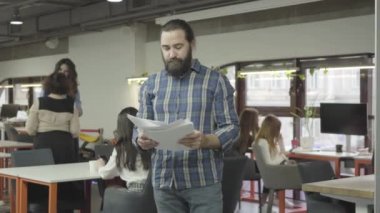 Modern ofiste ayakta kağıt belgeleri okuyan ciddi sakallı adam konsantre. İki kadın arka planda konuşuyor. Ofiste aktif çalışma ömrü