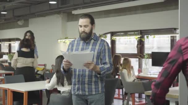 Ambiente de trabajo en una oficina respetable. Un oficinista monta una bicicleta alrededor de la oficina y derriba a un colega barbudo de pie con documentos en el lugar de trabajo . — Vídeos de Stock