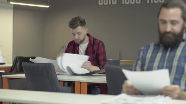 Ofiste oturan iki iş arkadaşı gazetelerden yeni çalışma bilgileri okuyor. Arka planda genç adam sakallı eski iş arkadaşı tavsiye soran ama o tarafına kollarını yayılır — Stok video