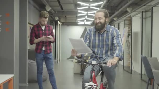 Barbudo sorrindo homem positivo anda de bicicleta mais perto com papéis na mão e mostra documentos para a senhora no escritório. Mulher bonita e homem barbudo se comunicam no escritório, discutindo novo documento — Vídeo de Stock