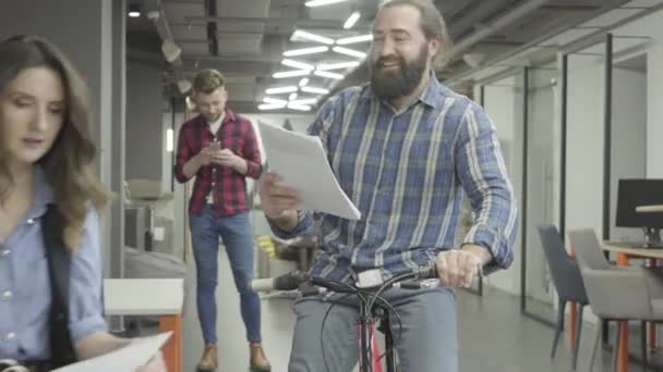 Skäggiga leende mannen rider sin cykel med papper i hand och visar dokument till damen på kontoret. Vacker kvinna och skäggiga man kommunicerar på kontoret, diskuterar nya dokument, de är glada — Stockvideo