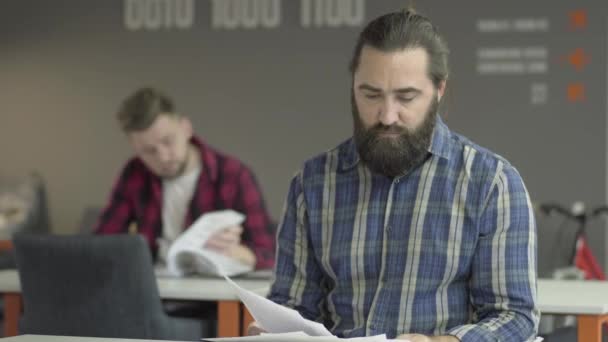 Hombre barbudo enojado sentado en primer plano estudiando papeles en la oficina. El trabajador se vuelve hacia su colega y arroja papeles a su mesa, luego se levanta y se aleja. Problema en el proceso de trabajo — Vídeos de Stock