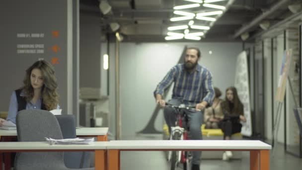 Barbudo sonriente hombre monta su bicicleta en la oficina moderna. Rider saluda a su colega y se sienta en su lugar de trabajo. Trabajador de oficina llegó al trabajo en la bicicleta. El hombre comienza su día de trabajo — Vídeo de stock