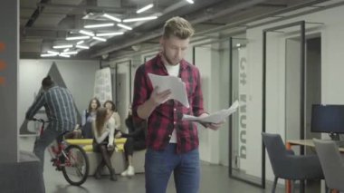 Modern ofiste ayakta kağıt belgeleri okuyan düşünceli sarışın genç adam. Kadın meslektaşları arka planda sohbet. Motosiklete binmek sakallı adam. Ofiste aktif çalışma ömrü