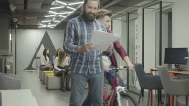 Ofiste gazete okuyan sakallı adam. Ofiste bisiklet sürme ve omzunda sakallı adam iter Genç meslektaşım, kağıtlar düşmek. Bisiklet binici özür dileyen ve belge toplama