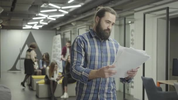 Hombre barbudo concentrado estudiando documentos de los papeles de pie en la oficina. Joven colega montando la bicicleta en la oficina y empuja al tipo barbudo en el hombro, los papeles se caen. Beardie está enojado. — Vídeo de stock