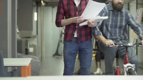 Bebaarde kantoormedewerker rijden een fiets op het kantoor. Een man klopt zijn collega staande met papieren op de werkplek en het bestuderen van documenten met verslagen en plannen voor de toekomst. Werkplek. — Stockvideo