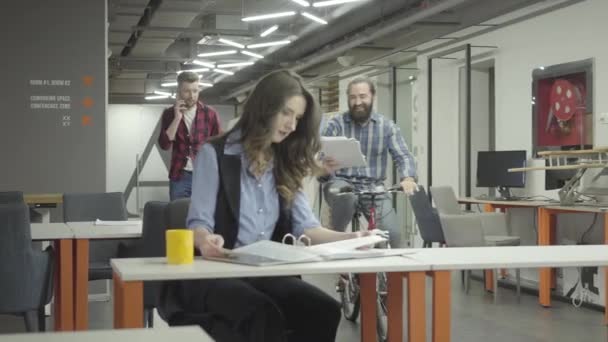 Mujer bonita sentada en su lugar de trabajo en la oficina haciendo su trabajo. Barbudo sonriente hombre monta su bicicleta más cerca con papeles en la mano y muestra documentos a la señora. Colegas sonriendo — Vídeos de Stock