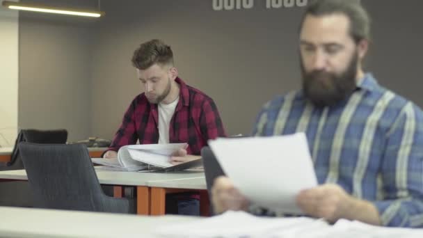 Двоє бородатих офісних працівників працюють в офісі, вивчаючи звітну документацію. Колеги читають документи з робочими планами. Один офісний працівник або бізнесмен обурений поганими покачанами і криками — стокове відео