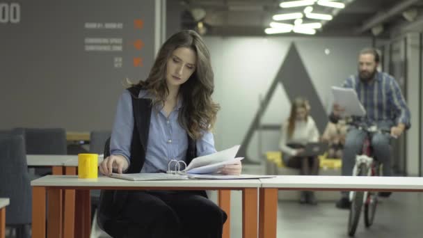 Bella donna seduta sul posto di lavoro in ufficio a fare il suo lavoro. Uomo positivo barbuto guida la sua moto più vicino con carte in mano e mostra i documenti alla signora. Donna sorridente, ha letto buone notizie — Video Stock