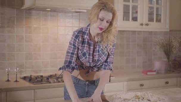 Retrato linda chica apasionada con un cuerpo sensual rueda la masa con un rodillo mientras está de pie en la mesa de la cocina. Chica seductora en una camisa a cuadros con vientre desnudo . — Vídeos de Stock