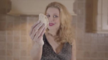 Büyük göğüsleri ile genç sarışın kadın kamera Dumpling göstermek ve sonra parçalara hamur ripping. Evde mutfakta hamur hazırlama tutkulu kız.