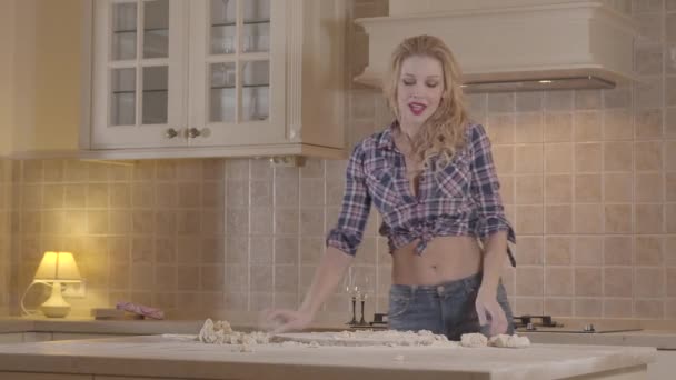 Uwodzicielska dziewczyna z zmysłowym ciałem przygotowuje ciasto stojąc przy stole kuchennym. Wesoła dziewczyna ugniatanie ciasta i wieje mąkę z toczenia PIN. — Wideo stockowe