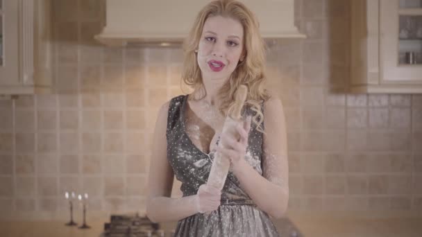 Porträtt ung smiley blond kvinna med rött läpp stift och stora bröst i svart klänning gnuggar brödkavel med mjöl. Passionerad flicka förbereder degen i köket hemma. — Stockvideo