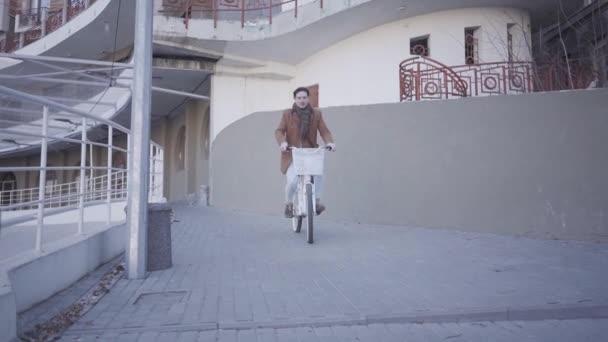Szczęśliwy uśmiechnięty człowiek w brązowym płaszczu jazda na rowerze w mieście w pobliżu dużego budynku. Wypoczynek z przystojnym miastem mieszkań. Aktywny styl życia — Wideo stockowe