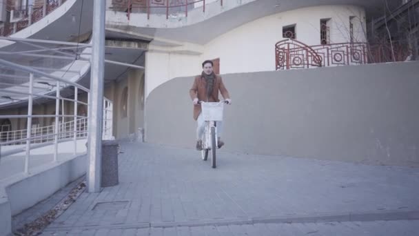 Happy smiling man in brown coat riding his bicycle in the city near large building. Leisure of handsome city dweller. Active lifestyle — Stock Video