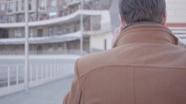 Cep telefonu ile konuşan şehir Caddesi üzerinde yürümek kahverengi ceket yakışıklı emin adamın arka görünümü. Arka planda kentsel şehir manzarası. Modern şehir sakinleri. Kamera adam takip