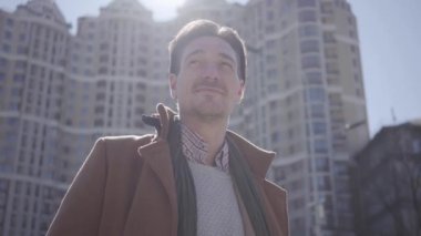 Şehir sokakta görünümlü kahverengi ceket ayakta gülümseyen yakışıklı emin adam portresi. Arka planda yüksek binalar. Alttan çekim