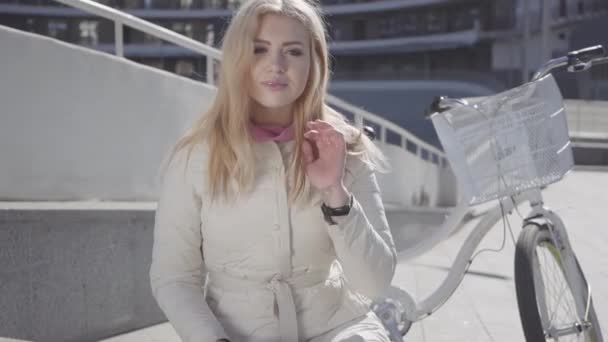 Mujer rubia bonita con chaqueta blanca cálida sentada en la calle de la ciudad con bicicleta detrás mirando a la cámara. Ocio de bonito habitante de la ciudad. Estilo de vida activo. Movimiento lento — Vídeo de stock