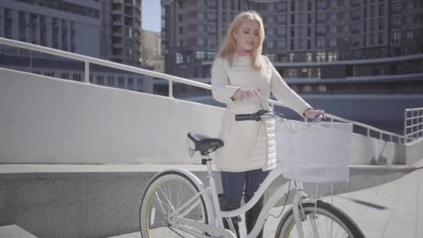 Mujer rubia bonita con chaqueta blanca cálida de pie en la calle de la ciudad con la bicicleta mirando a la cámara tocando su cabello. Ocio del joven habitante de la ciudad. Estilo de vida activo. Movimiento lento — Vídeos de Stock