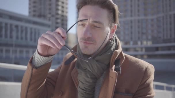 Portrait d'un bel homme en manteau marron debout dans la rue de la ville portant des lunettes de soleil. Loisirs du citadin. Paysage urbain en arrière-plan. Mouvement lent . — Video