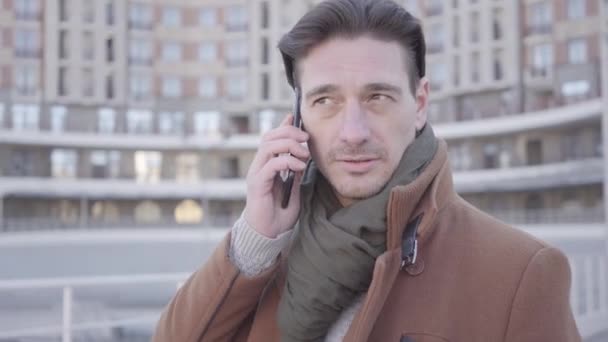 Portræt af flot selvsikker mand i brun frakke stående i byen gaden taler via mobiltelefon. Byernes bybillede i baggrunden. Moderne byboer – Stock-video
