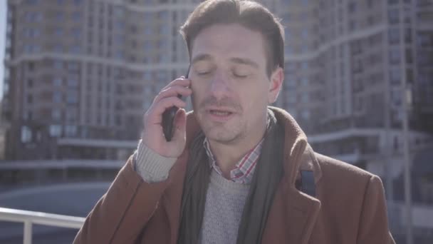 Retrato de un hombre guapo y confiado en abrigo marrón parado en la calle de la ciudad hablando por teléfono celular. Paisaje urbano en el fondo. Habitante de la ciudad moderna. Cámara se mueve alrededor — Vídeo de stock