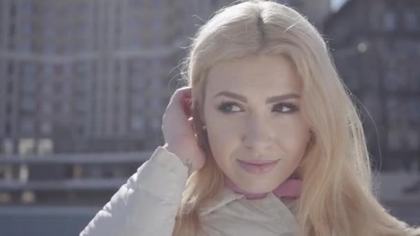 Retrato de mulher loira bonita correndo a mão através de seu cabelo e olhando para a câmera sorrindo. A paisagem urbana ao fundo. Habitante da cidade moderna — Vídeo de Stock