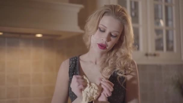 Blonde vrouw met grote borsten in zwarte jurk kneed het deeg in de handen op de keuken — Stockvideo