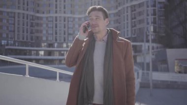 Şehir sokakta cep telefonu ile konuşan kahverengi ceket ayakta yakışıklı emin adam portre. Arka planda kentsel şehir manzarası. Modern şehir sakinleri. Kamera etrafında hareket eder