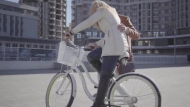 Handsome man in brown coat teaching his girlfriend to ride the bicycle in the city, both people laughing. Leisure of beautiful couple of city dwellers. Active lifestyle