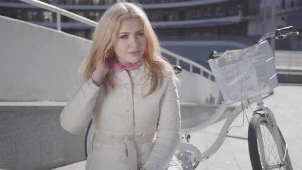 Portret van een jong mooi meisje met blond haar en in een licht jasje zittend op een bankje naast de fiets. Het meisje is te typen op een cel in goed weer buitenshuis. — Stockvideo